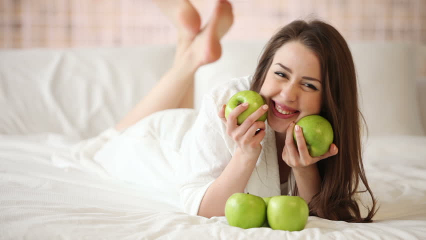 Cheerful young woman lying in bed with apples in front of her holding two of