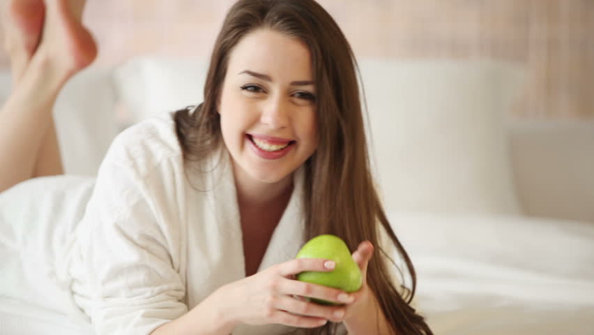 Charming young woman relaxing in bed holding green apple looking at camera and
