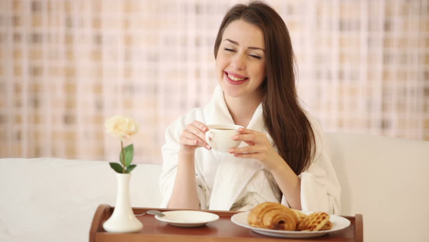 Cute girl in bathrobe sitting in bed drinking tea looking at camera and smiling.