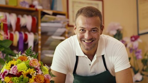 mid adult man working as florist in flower shop and looking at camera, smiling with bouquet on desk