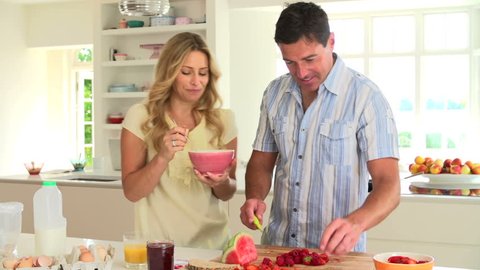 Man cuts fresh fruit for brunch while woman eats breakfast cereal and drinks orange juice