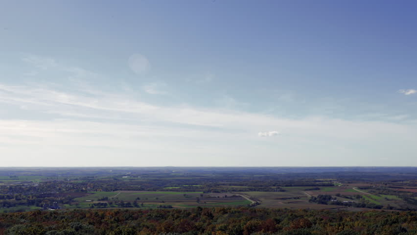 Timelapse - wide view of Wisconsin countryside in the Fall. Shot from the Blue Mounds State Park hill, clouds soar over treetops bright with fall colors, green fields, farmhouses and rural homesteads.