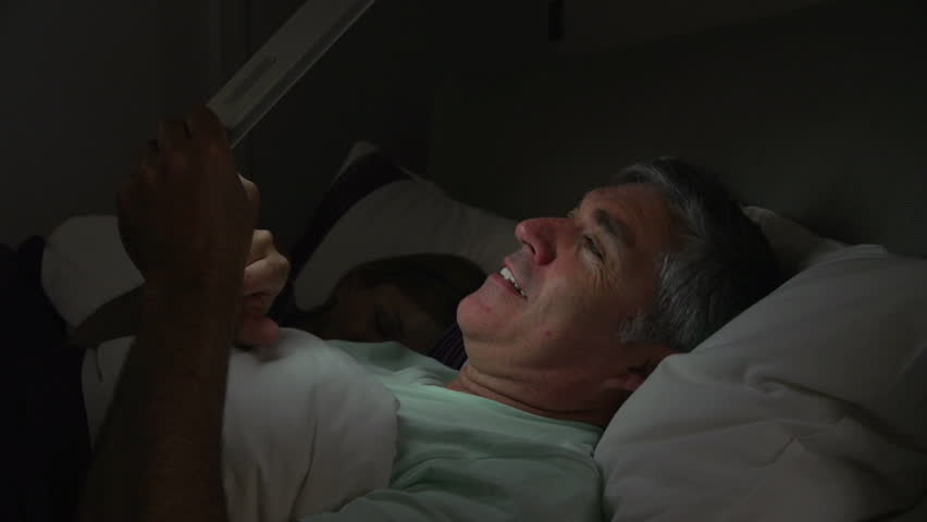 Middle aged man lies in bed with face illuminated by digital tablet while wife sleeps