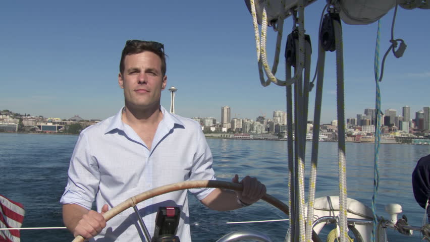Man takes the helm of a sailboat with the city skyline behind him.  Filmed on