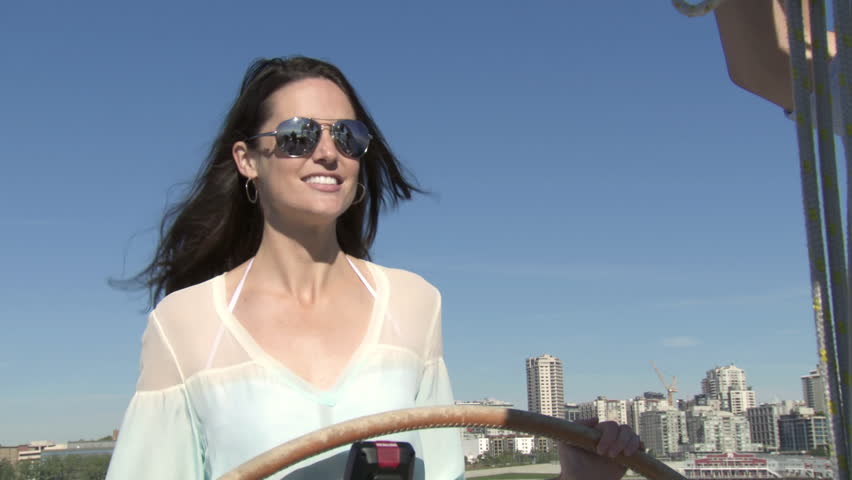 Attractive woman takes the helm of a sailboat with the city skyline behind her. 