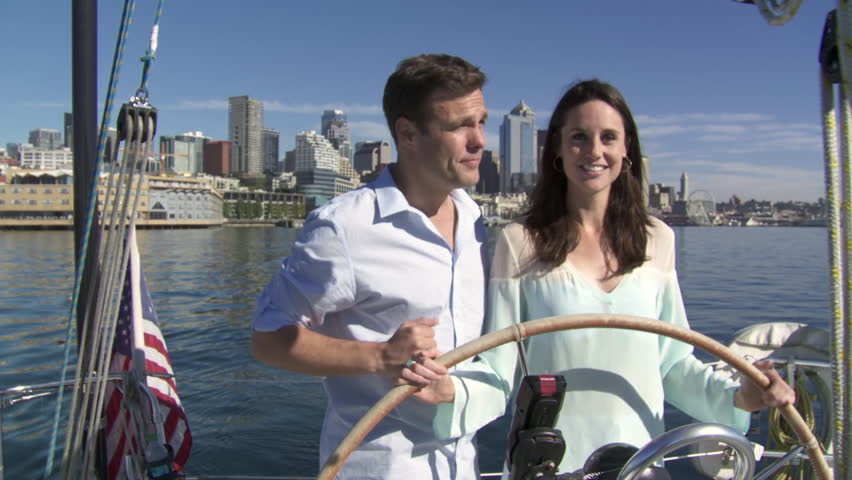 Young couple takes the helm of a sailboat with the city skyline behind them. 