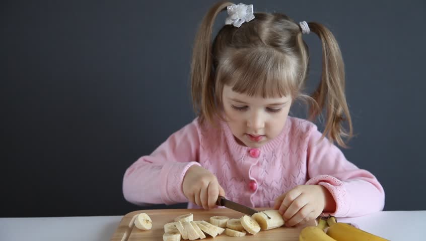 Charming little girl cutting a ripe banana and showing thumb up, grey background