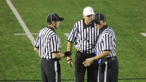 Three football referees stand in a group and discuss a call. The head ref signals pass interference as the penalty