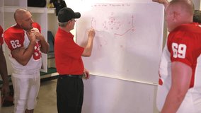 Football players study the plays their coach is drawing out on a whiteboard in the locker room before a game