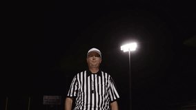 A football referee makes a false start penalty hand signal in front of stadium floodlights