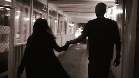 A couple holds hands as they walk down a tunnel at night in the city. Black and white shot
