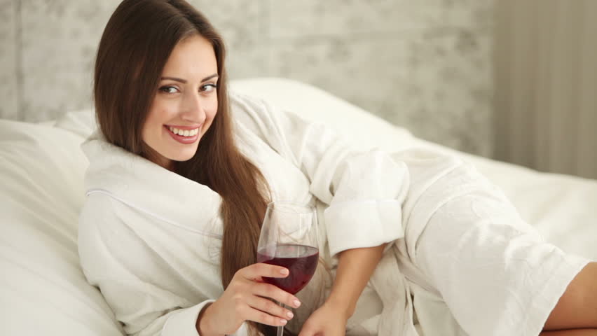 Attractive girl lying in bed holding glass of wine looking at camera and