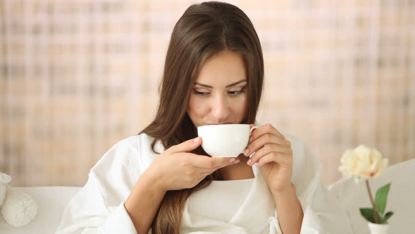 Attractive young woman relaxing in bed drinking tea looking at camera and