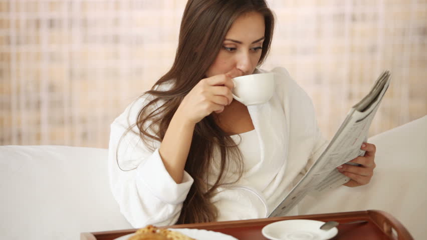 Beautiful girl relaxing in bed reading newspaper drinking tea and smiling.