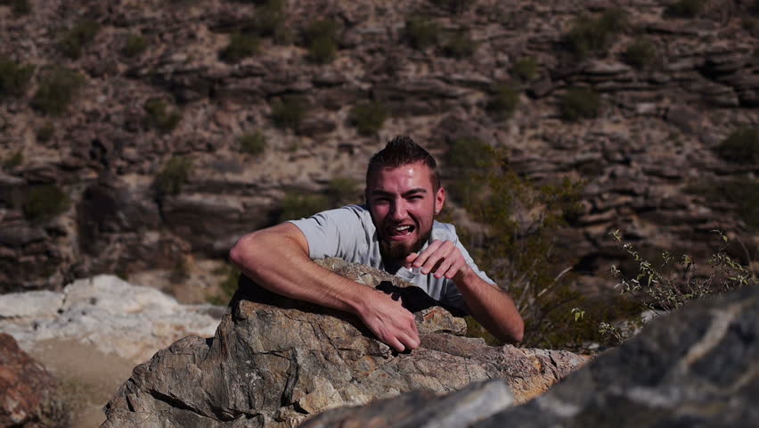 A young man climbs up a cliff in the desert then falls away.