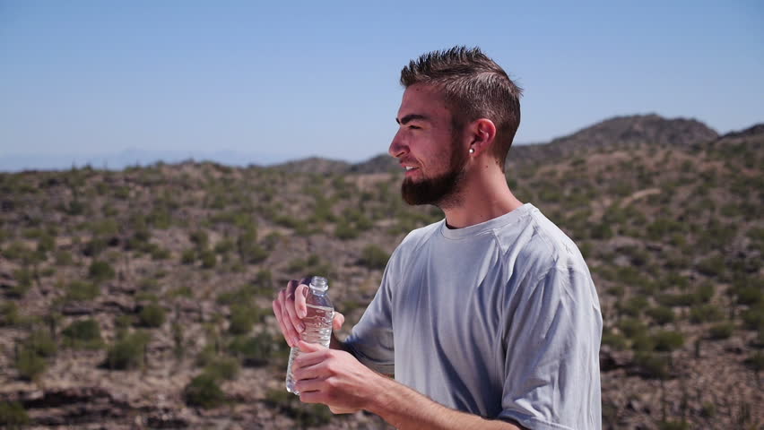 A young man hikes though the desert and takes a drink of water from a plastic