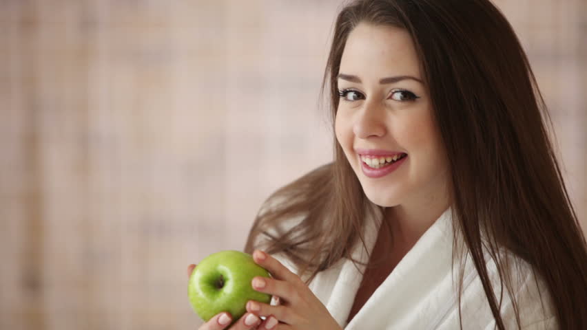 Charming girl sitting on bed holding green apple looking at camera and smiling.