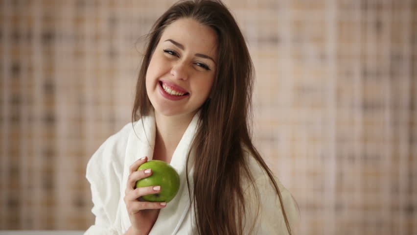 Beautiful girl sitting on bed holding green apple looking at camera and smiling.
