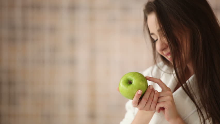 Attractive girl sitting on bed holding green apple looking at camera and