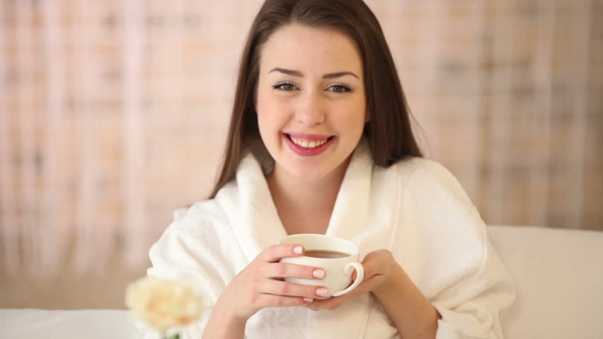Beautiful girl sitting in bed drinking tea looking at camera and smiling.