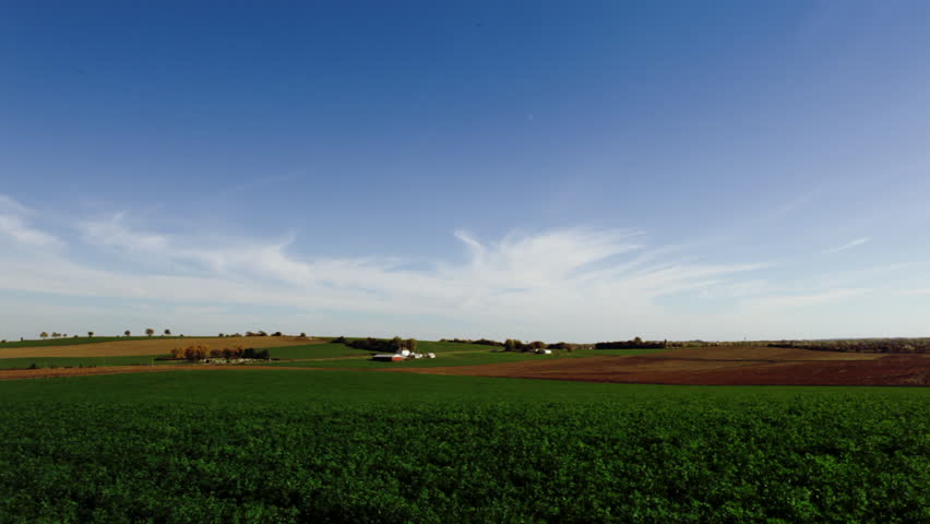 Wisconsin farmland, a green field in the foreground, blue sky with wispy clouds overhead. Widest view.
