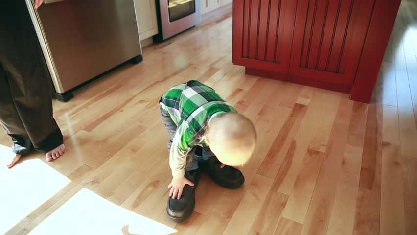 A little boy walking around the kitchen with his mother in his fathers shoes