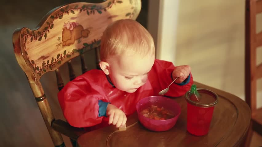 A little baby boy eating stew in his high chair in the kitchen at his home