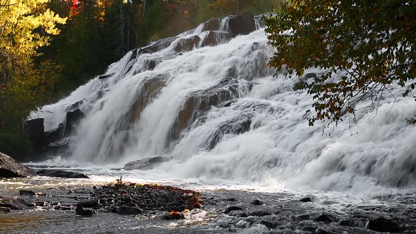Whitewater cascades over boulders with yellow autumn foliage and golden morning light in the mist of  Bond Falls, a waterfall in Michigan