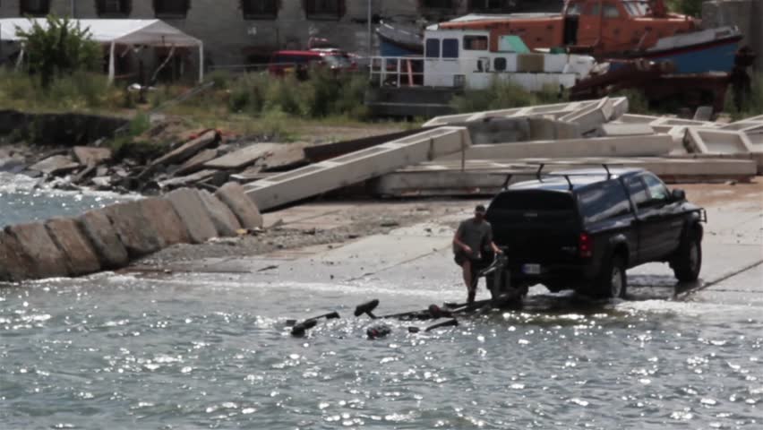 Speed boat docking at shore where a black family truck is waiting for the boat to dock in the carrier found at the back of the truck. 
