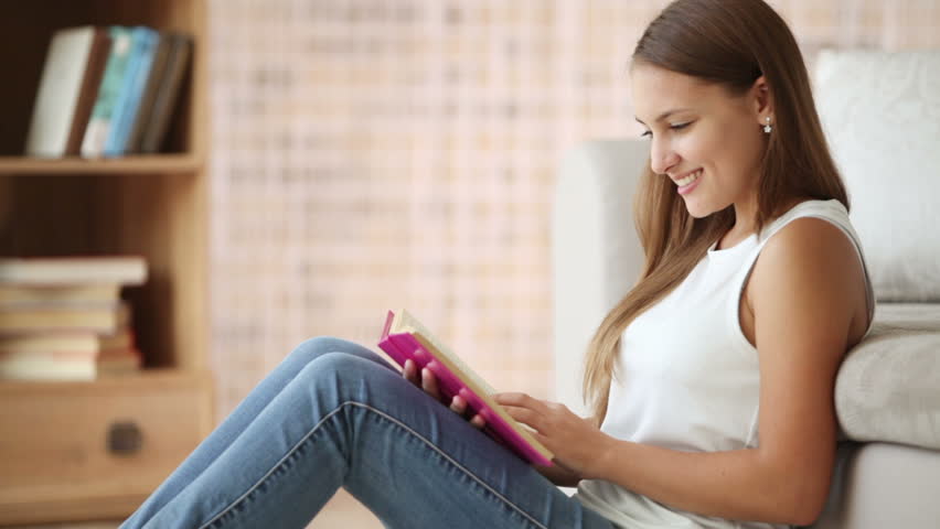 Attractive girl sitting on floor by sofa reading book and smiling. Panning
