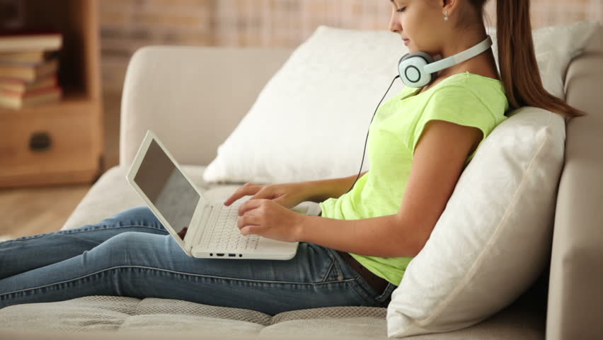 Attractive girl in headset sitting on sofa using laptop and smiling at camera.