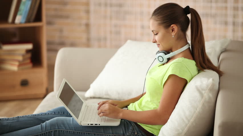 Beautiful girl in headset sitting on sofa using laptop looking at camera and