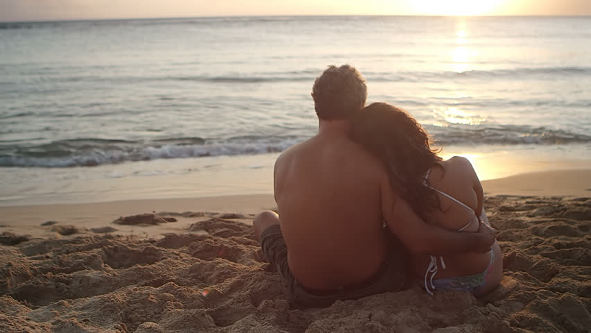 A couple sits in the sand on the beach, hugging, watching the sun set over the ocean