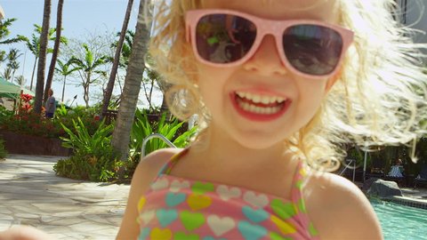 A young girl wearing sunglasses at the pool smiles and laughs at the camera
