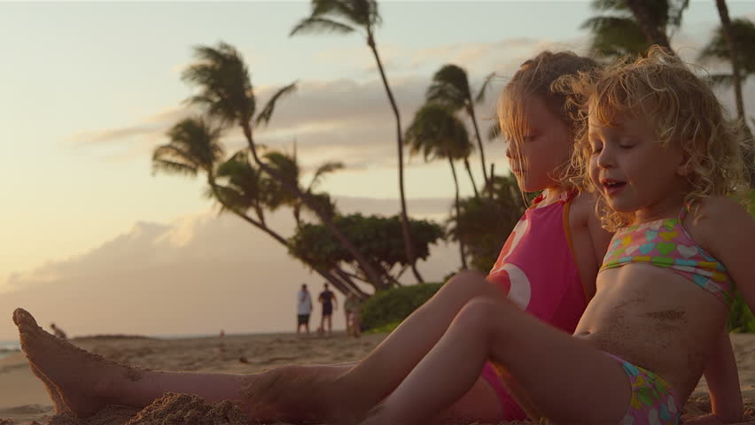A cute little girl hugs her sisters as they sit on the beach during sun set. Medium shot.