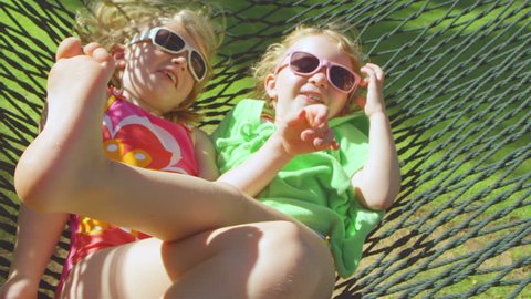 Two adorable little girls lie on a hammock together as it swings back and forth. Medium shot.
