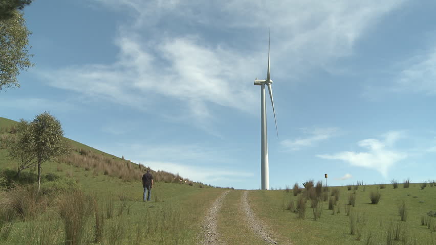 A man walks towards a tall wind turbine in a countryside field