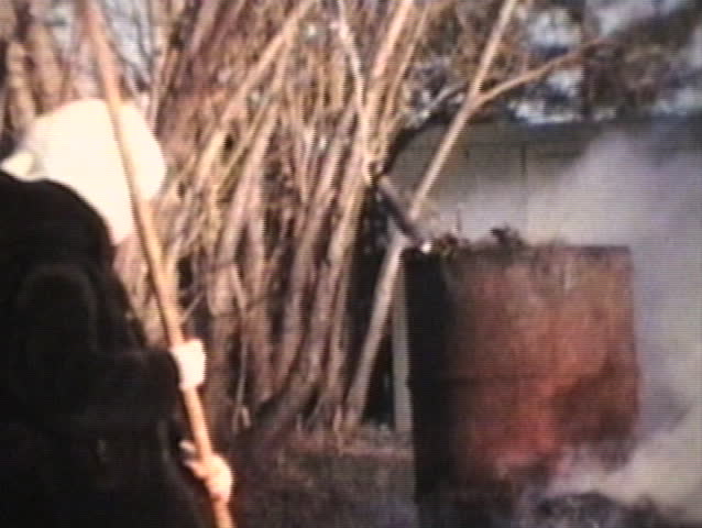A young girl rakes leaves and puts them on the burn pile in the backyard. Also in the shot is an old oil drum with smoke pouring out of it as well.