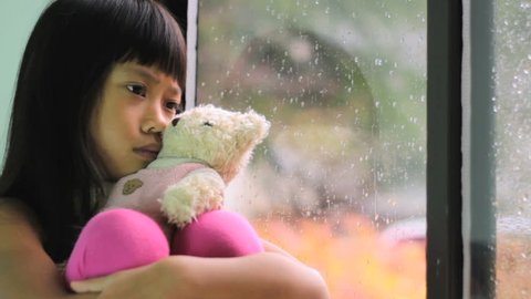 A sad little seven year old Asian girl sits by the window with her Teddy Bear watching the rain drops fall.
