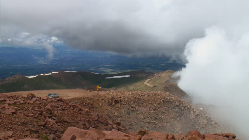 Looking north from summit of Pikes Peak as cars wind their way down the road past cloud banks rapidly rising against the mountain.