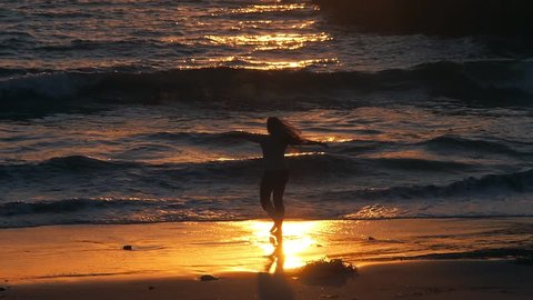 A medium shot of a girl spinning and playing in the beach waves at Playa Del Ray, Los Angeles, in an orange and yellow sunset on a blue ocean next to a pier., videoclip de stoc