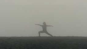 A girl with long hair does yoga, warrior pose, on a very foggy beach at Playa Del Ray, Los Angeles, in silhouette.