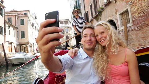 Couple in Venice on Gondole ride romance in boat happy together on travel vacation holidays. Romantic young beautiful couple taking self-portrait sailing in venetian canal in gondola. Italy.