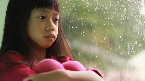 A depressed little seven year old Asian girl sits by a rainy window.