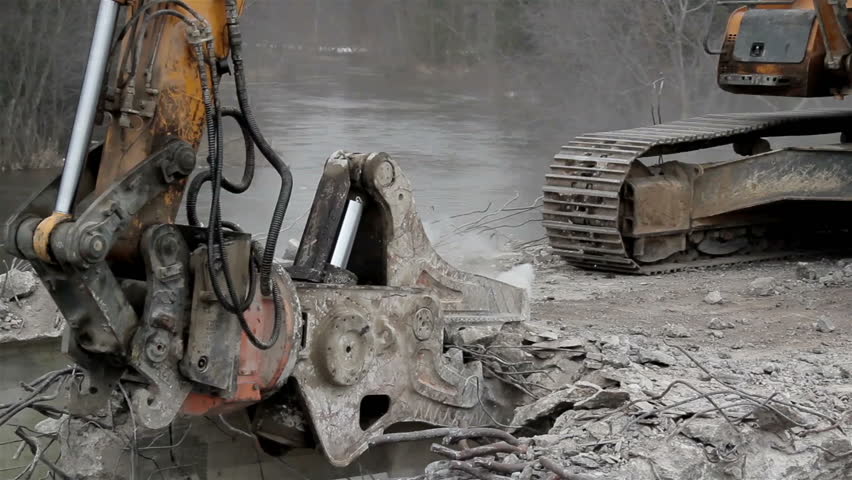 A bulldozer destroying a bridge into pieces; getting out the cement and the iron rods. Heavy equipment with pincher removing damaged bridge and demolish it completely before building a new one.