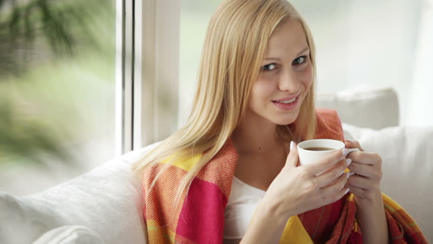 Cute young woman sitting on sofa drinking tea from cup looking at camera and