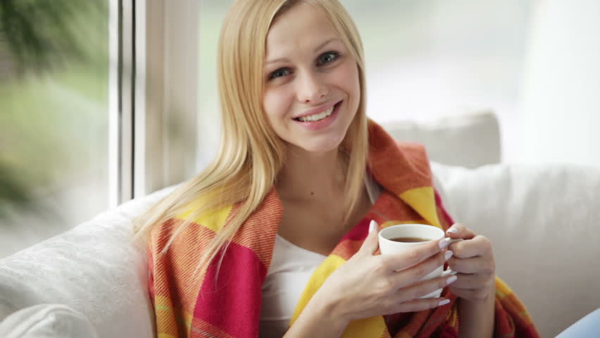 Young woman sitting on sofa drinking tea from cup looking at camera and smiling.