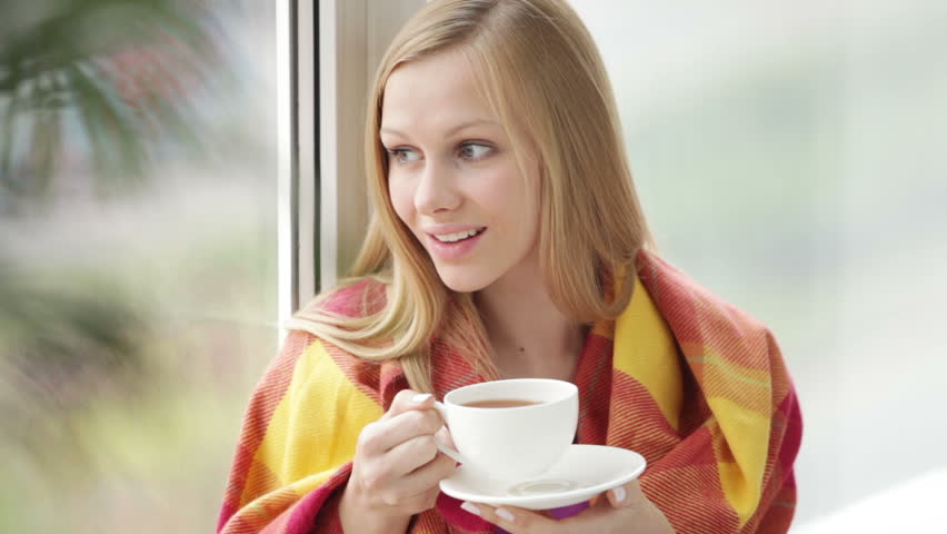 Pretty girl sitting by window drinking tea looking at camera and smiling.