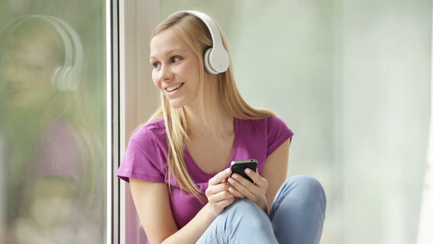 Pretty girl in headset sitting by window using cellphone looking at camera
