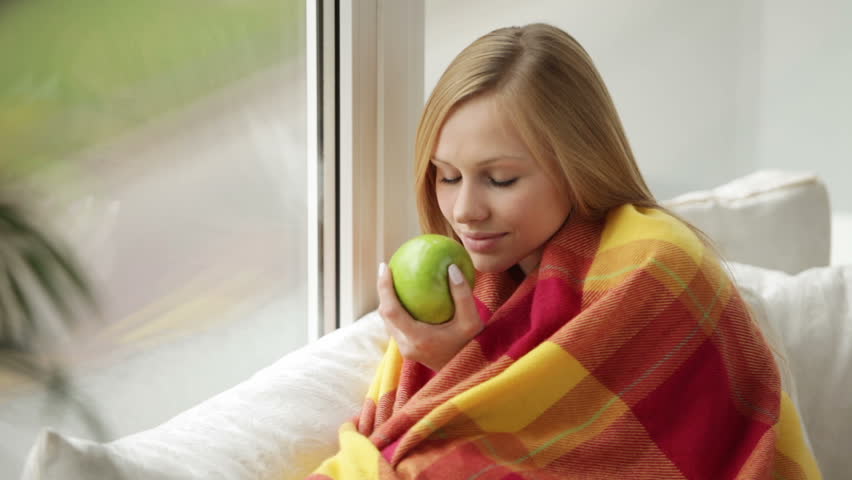 Cute young woman sitting on sofa holding apple looking out of window and smiling
